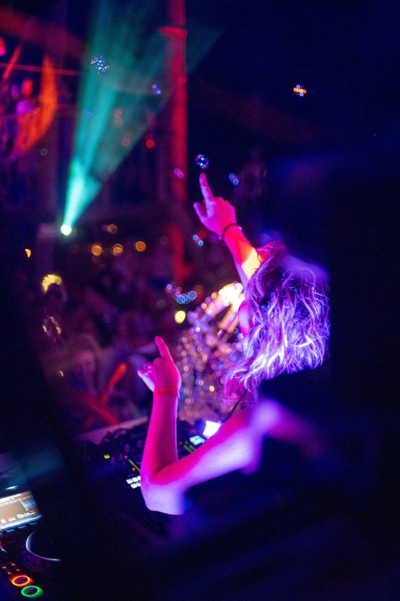 Daniela at the decks, seen from behind, pointing up into lasers and drifting bubbles above the crowd