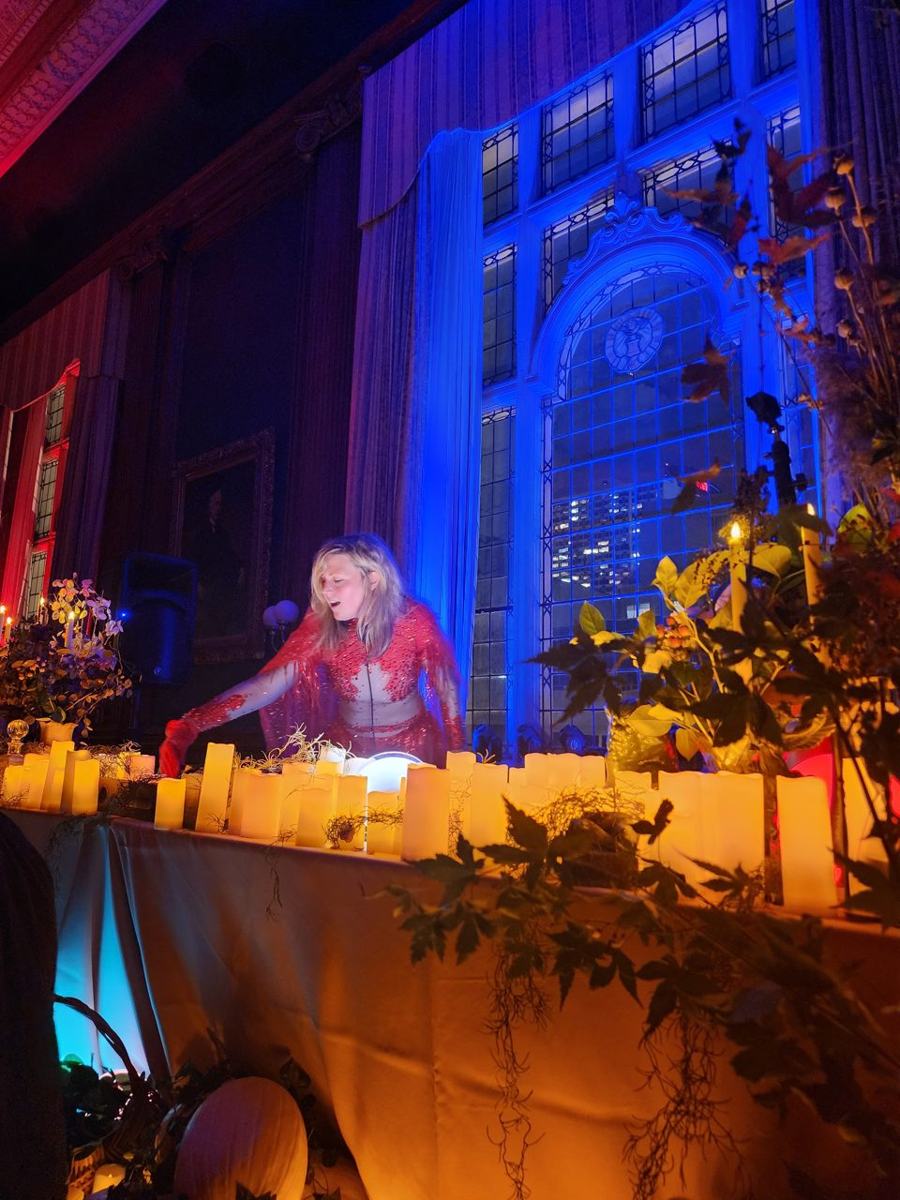 Daniela at the decks in red behind a bank of candles and foliage