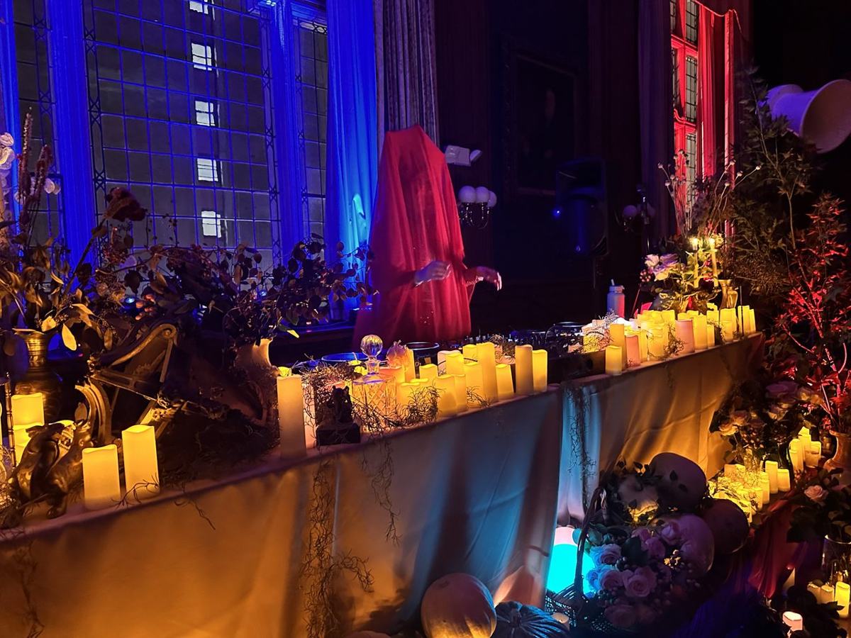 A figure veiled in red behind a long table of candles and flowers, blue-lit windows behind